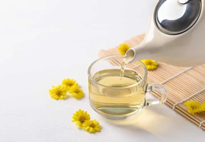 Tè giallo dorato versato da una teiera in una tazza trasparente, circondato da piccoli crisantemi fiori gialli.