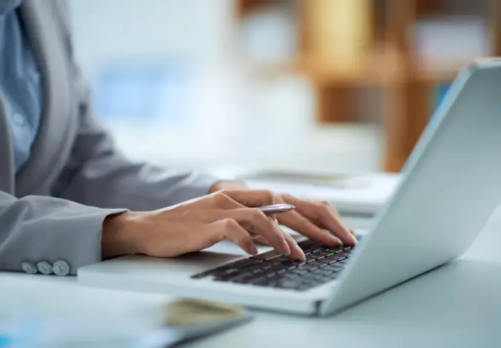 Dłonie piszące na klawiaturze laptopa, w tle biuro.
