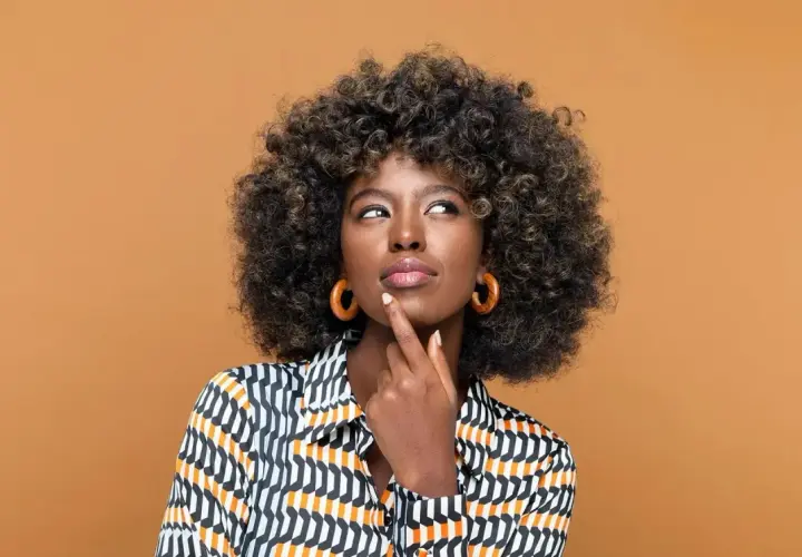 Mujer pensativa con cabello rizado y coloración semipermanente, luciendo una blusa estampada y aretes circulares.