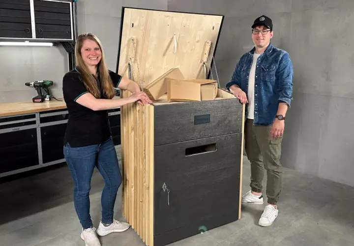Ein Paar präsentiert stolz seine selbstgebaute Paketbox. Die Box ist aus Holz gefertigt und hat eine Klappe, die mit Ketten gesichert ist. Sie ist groß genug, um mehrere Pakete aufzunehmen.