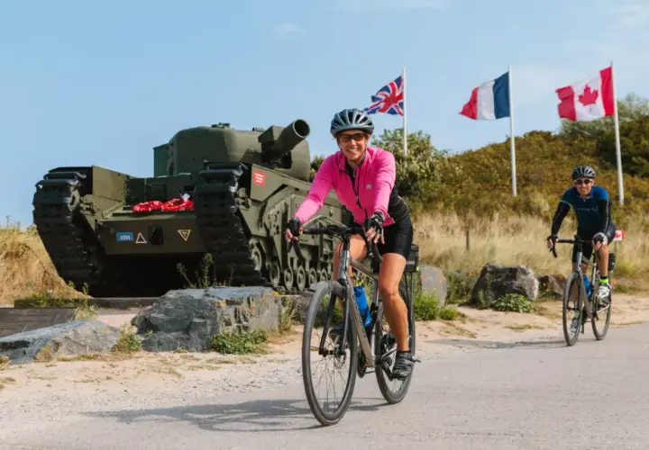 Cyclists enjoy Normandy bike tours past a tank memorial with Union Jack, French, and Canadian flags flying.