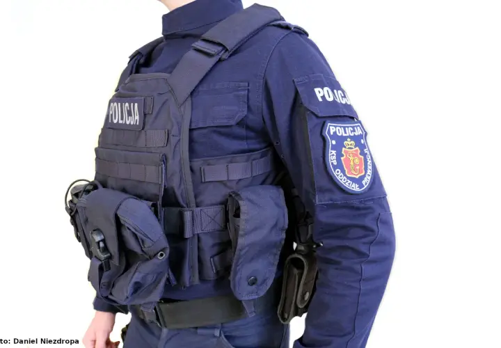Policjant w pełnym rynsztunku, z kamizelką kuloodporną z napisem "POLICJA" i naszywką "KSP ODDZIAŁ PREWENCJI".