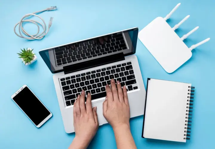 Ręce na klawiaturze laptopa, obok router wifi, telefon i kabel. Szukasz sposobu, jak zwiększyć zasięg wifi w laptopie?