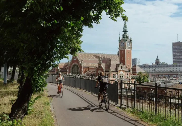 Dwie osoby na rowerach jadą ścieżką obok torów kolejowych. W tle piękny budynek dworca z wieżą zegarową. To idealne miejsce przyjazne rowerzystom.