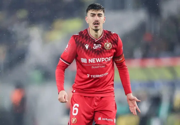 Piłkarz w czerwonej koszulce z numerem 6, z opaską na ramieniu z napisem "Widzew Łódź". W tle widać fragment stadionu, być może podczas meczu z Cracovią.