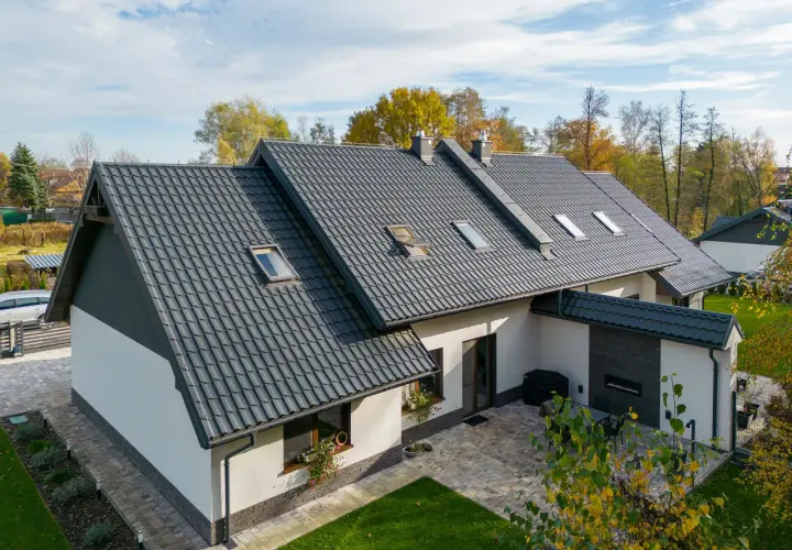 Nowoczesny dom z białymi ścianami i ciemnym, eleganckim dachem z dachówki ceramicznej. Wokół zieleń i jesienne drzewa.