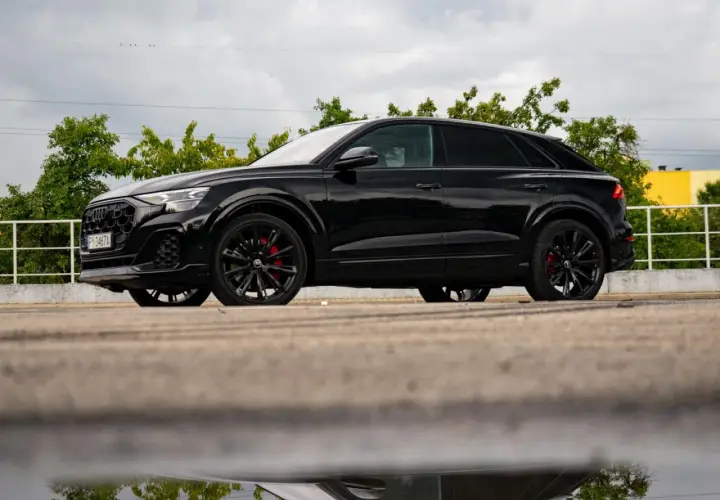 Czarny Audi Q9 z czerwonymi zaciskami hamulcowymi, odbijający się w kałuży. Sprawdź audi q9 cena!