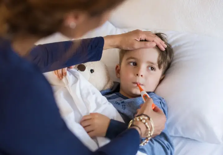 Chłopiec leży w łóżku z termometrem w ustach, mama sprawdza mu czoło. Widać, że jest w stanie podgorączkowym.