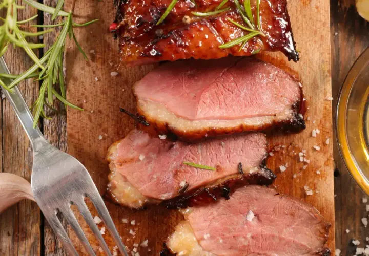 Magret de canard rosé, tranché sur une planche en bois, garni de romarin. Une fourchette et une gousse d'ail sont à proximité. Un délice pour accompagner un bon vin.