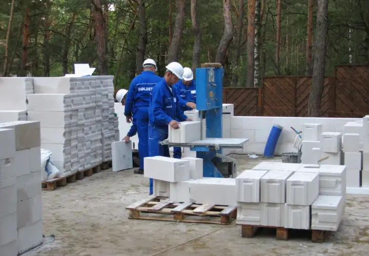 Na budowie pracownicy w niebieskich kombinezonach i kaskach układają bloczki betonowe. Widać kilka palet z bloczkami, gotowych do użycia.