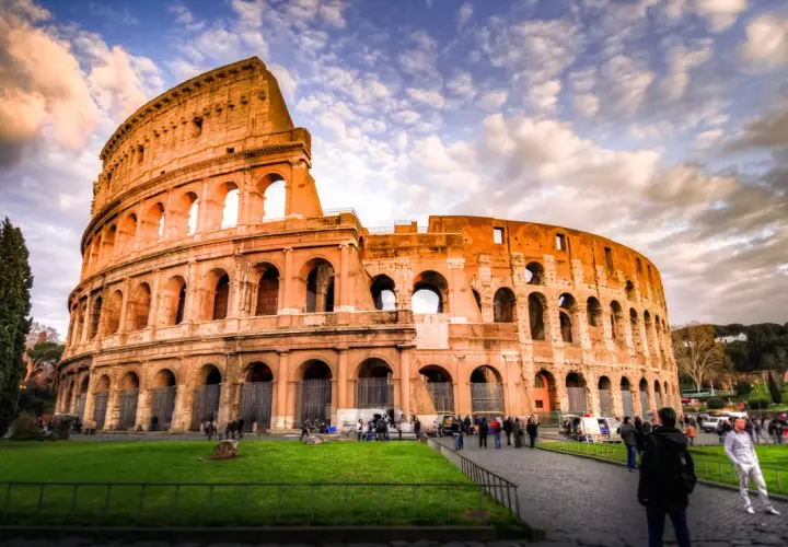 Il Colosseo, un'icona di cosa visitare a Roma, si erge maestoso sotto un cielo drammatico. Turisti passeggiano sul selciato, ammirando l'antica grandezza.