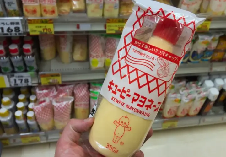 A hand holds a large squeeze bottle of Kewpie mayonnaise. The packaging features the iconic Kewpie doll. The question "how long does Kewpie mayo last?" is on my mind as I shop.
