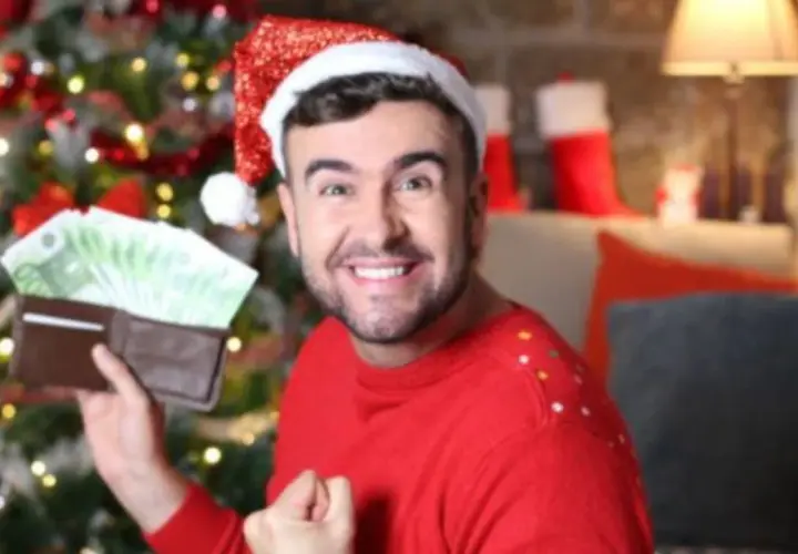 Hombre con gorro de Papá Noel muestra billetera llena de billetes, como si soñara que le toca la lotería en Navidad.
