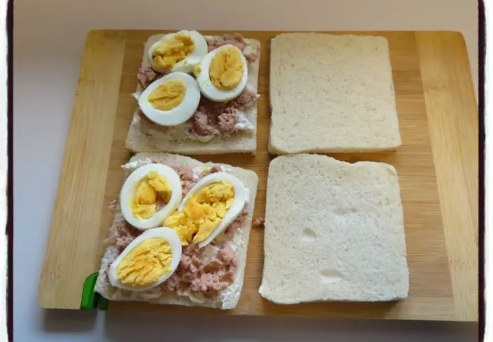 Deux sandwichs oeuf dur sont en préparation sur une planche en bois. Le thon émietté et la mayonnaise sont déjà étalés sur le pain.