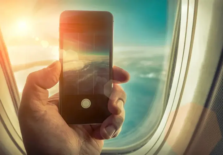Dłoń trzyma smartfon, którym fotografuje chmury z okna samolotu. Czy w samolocie można używać telefonu? Tak, do robienia zdjęć!