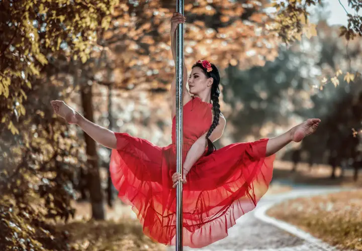 Kobieta w czerwonej sukience wykonuje figurę na słupie, jak podczas kursu pole dance. Jej nogi są rozłożone w szpagacie, a włosy splecione w warkocz.