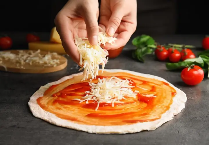Hände streuen geriebenen Gouda auf eine mit Tomatensauce bestrichene Pizza. Tomaten und Basilikum liegen daneben.