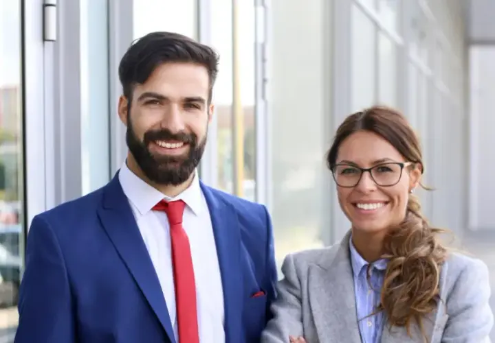 Smiling professionals, a man in a blue suit and red tie, and a woman in a grey blazer, stand outside. They're ready to pursue online MPA programs no GRE.
