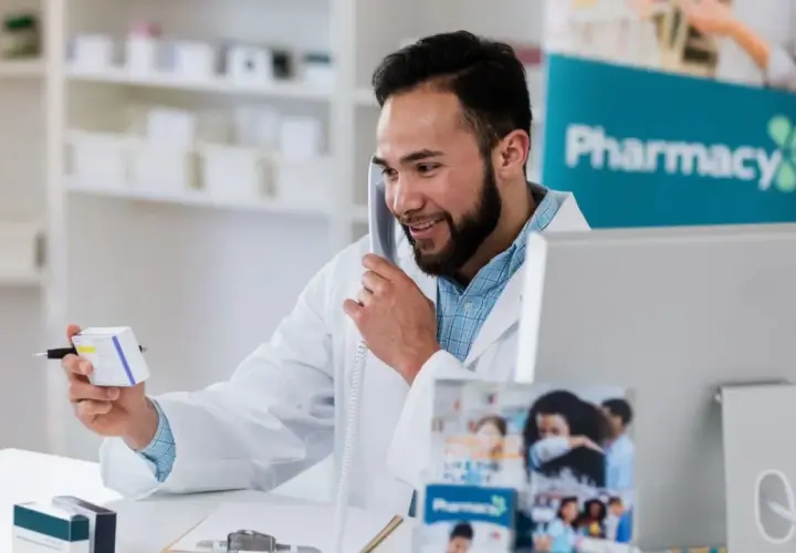 Uśmiechnięty farmaceuta rozmawia przez telefon, trzymając pudełko leku. Zastanawia się, ile zarabia farmaceuta, pracując w aptece.