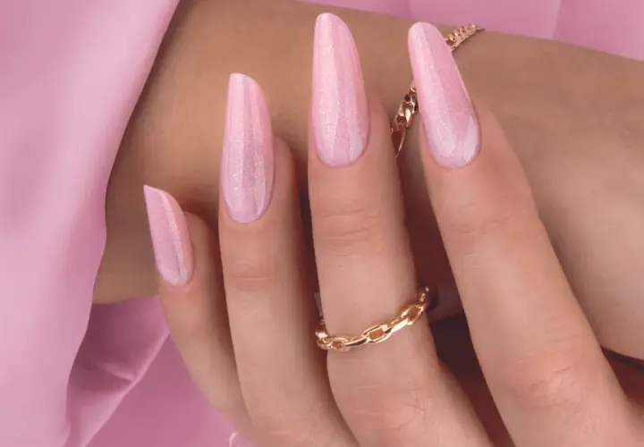 Delikatny, różowy manicure hybrydowy z subtelnymi drobinkami brokatu. Długie paznokcie w kształcie migdała, ozdobione złotą biżuterią.