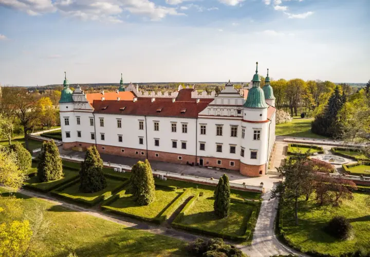 Zamek w Baranowie Sandomierskim, biała rezydencja z czerwonym dachem i zielonymi wieżyczkami, otoczona zadbanym ogrodem.