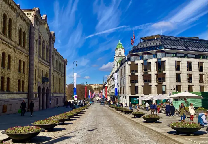 Słoneczny dzień na Karl Johans gate w Oslo. Brukowana ulica z rzędami kwietników prowadzi do Pałacu Królewskiego.