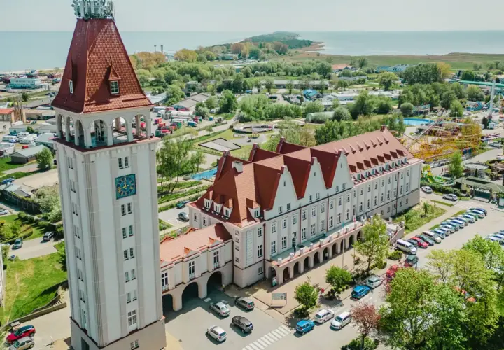 Wieża zegarowa i długi budynek z czerwonym dachem, przypominający dom rybaka, stoją obok siebie. W tle widać morze i park rozrywki.