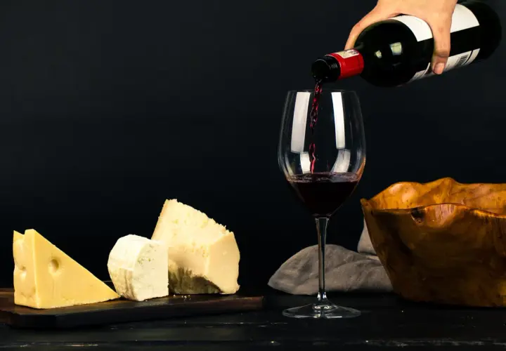 Un verre se remplit de vin rouge, à côté de morceaux de fromage et d'un bol en bois.