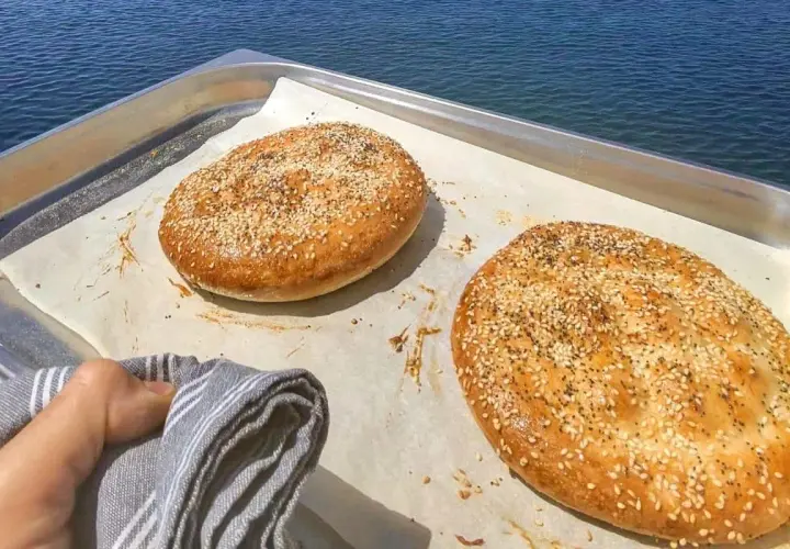 Två gyllene ramazan pidesi, toppade med sesamfrön, vilar på ett bakplåtspapper vid havet.