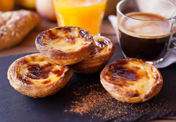 Pyszne babeczki portugalskie z kremowym nadzieniem i karmelizowaną skórką, obok kawy i soku pomarańczowego.