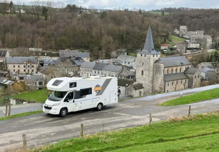Un camping-car blanc roule dans un village pittoresque de Belgique, avec une église en pierre et des maisons anciennes.