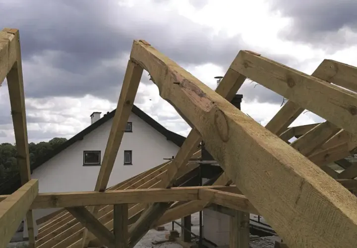 Montaż więźby dachowej w toku. Drewniane belki tworzą konstrukcję dachu nad białym domem z kominem.