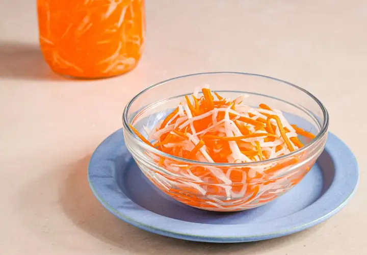 A bowl of vibrant japanese pickled carrots and daikon radish sits on a blue plate, with a jar of the same pickled vegetables in the background.