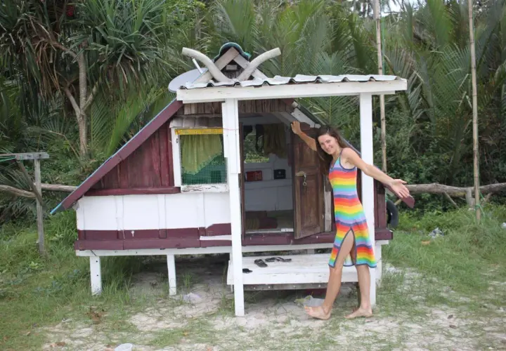 Uśmiechnięta kobieta w tęczowej sukience stoi przed małym domkiem na plaży. Idealne miejsce, gdzie najlepiej rezerwować noclegi, by poczuć tropikalny klimat.