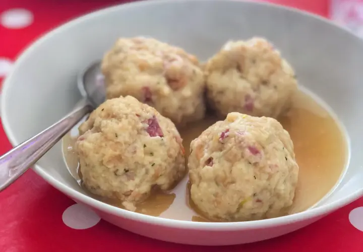 Canederli in brodo, un classico dei piatti tipici Alto Adige, serviti in una ciotola bianca su un tavolo rosso a pois bianchi.