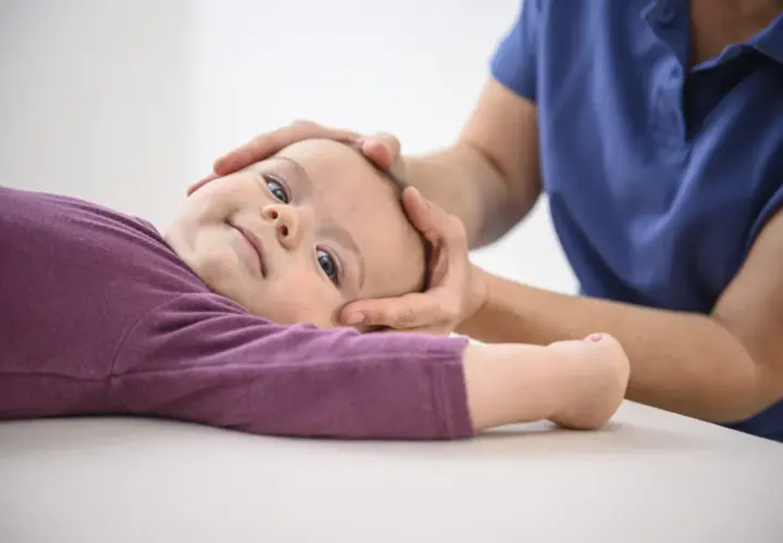Sanfte Hände einer Osteopathin untersuchen den Kopf eines lächelnden Kleinkindes.