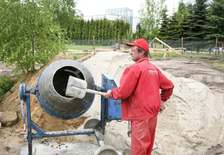 Budowlaniec w czerwonym ubraniu obsługuje betoniarkę na placu budowy, obok sterty piasku.