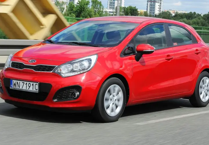 Czerwony Kia Rio 3 w ruchu, z widocznym numerem rejestracyjnym WN 7191C, na tle mostu i budynków.