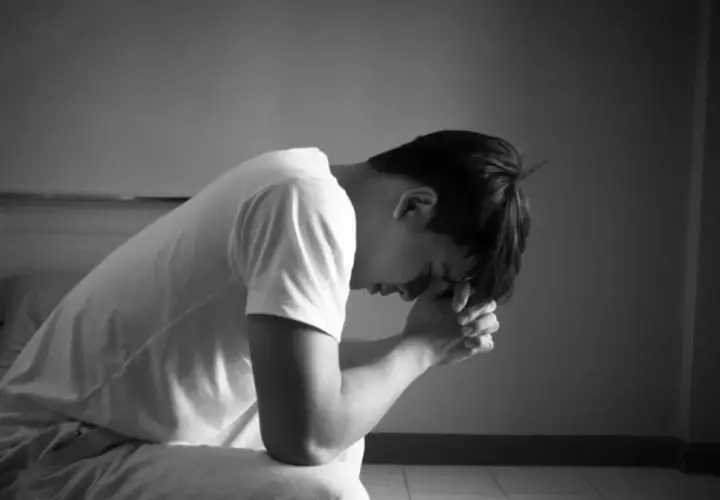 Mężczyzna siedzi na łóżku, pogrążony w melancholii, z rękami złożonymi przed twarzą.