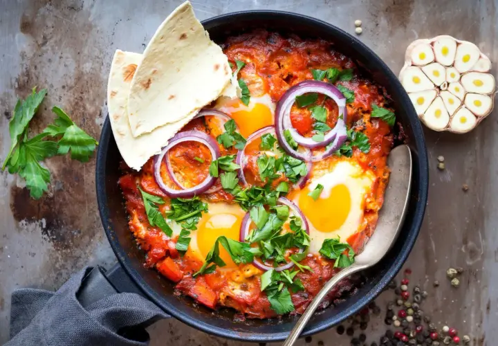 Délicieuse recette de chakchouka dans une poêle, garnie d'œufs, d'oignons rouges et de persil frais. Servi avec du pain pita.