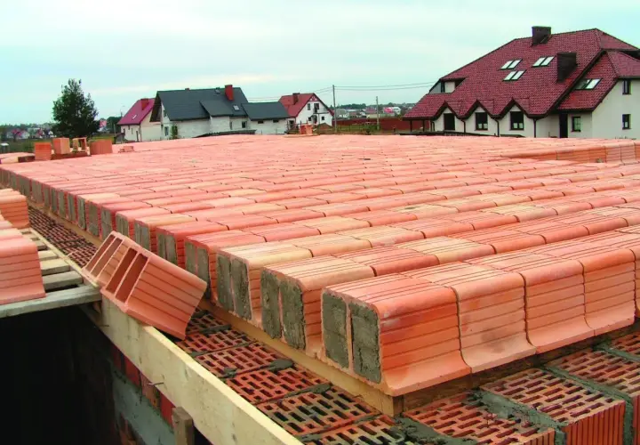 Budowa stropu ackermana. Czerwone pustaki ceramiczne ułożone w rzędach, tworzące konstrukcję stropu. W tle domy z czerwonymi dachami.