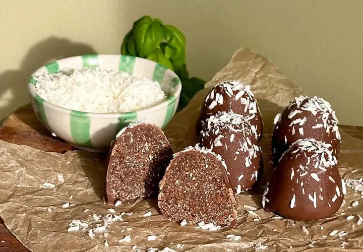 Hemmagjorda delicatobollar, täckta med choklad och kokosflingor, redo att avnjutas. En skål med kokosflingor står bredvid.
