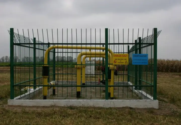 Ogrodzony obiekt gazowy z żółtymi rurami. Tablice ostrzegawcze informują o zagrożeniu i numerach alarmowych. Widać, że odległość budynku od gazociągu średniego ciśnienia jest bezpieczna.