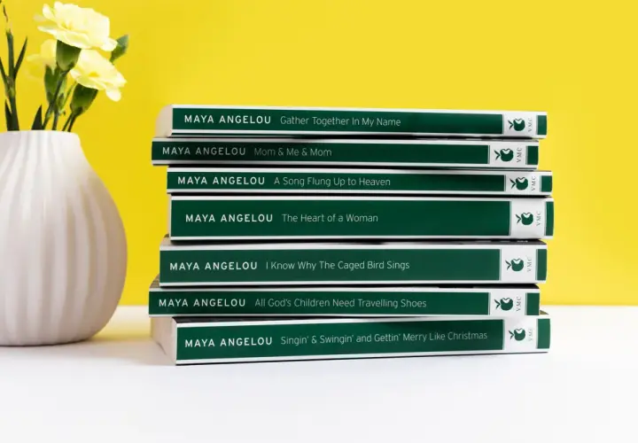 Stos **książek** **Maya Angelou** obok wazonu z żółtymi kwiatami.