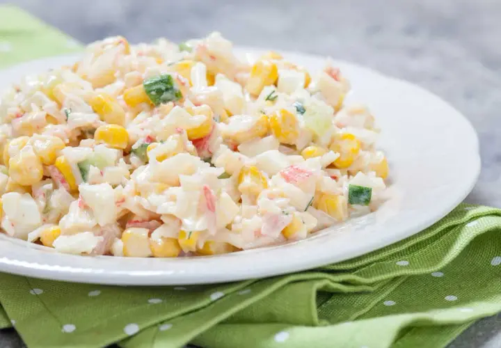 Deliciosa ensalada de arroz con mayonesa, maíz, pepino y surimi, servida en un plato blanco sobre una servilleta verde.