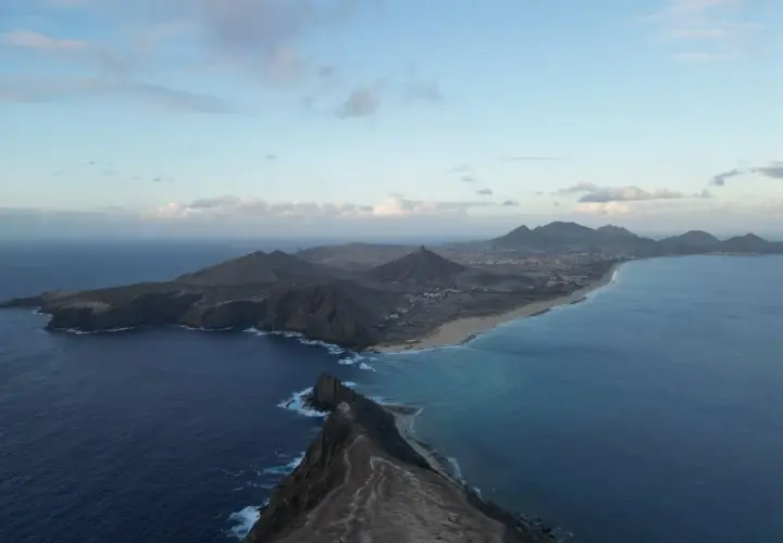 Dramatic coastline of Porto Santo island, with rugged cliffs meeting turquoise waters and a long sandy beach stretching towards distant mountains under a cloudy sky.