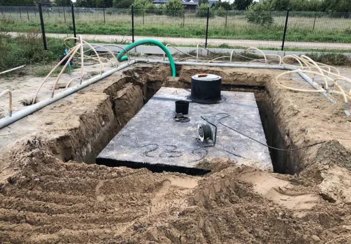 Widok na wykop pod szambo. Duża betonowa płyta, okrągły właz i liczne rury.
