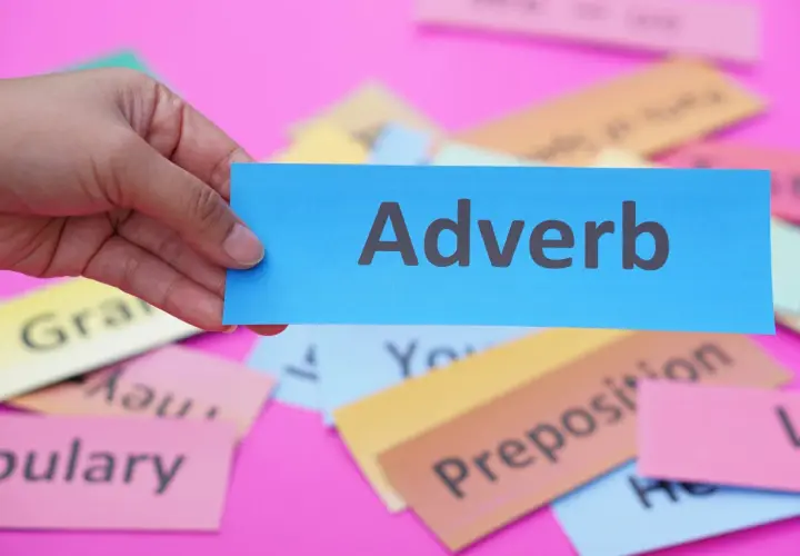 Una mano tiene un biglietto blu con la parola "Adverb". Sullo sfondo, altri biglietti colorati con parole come "Grammar" e "Vocabulary".