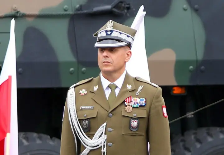 Oficer w mundurze, z orderami na piersi, stoi obok polskiej flagi. Nad nim widać fragment pojazdu wojskowego.