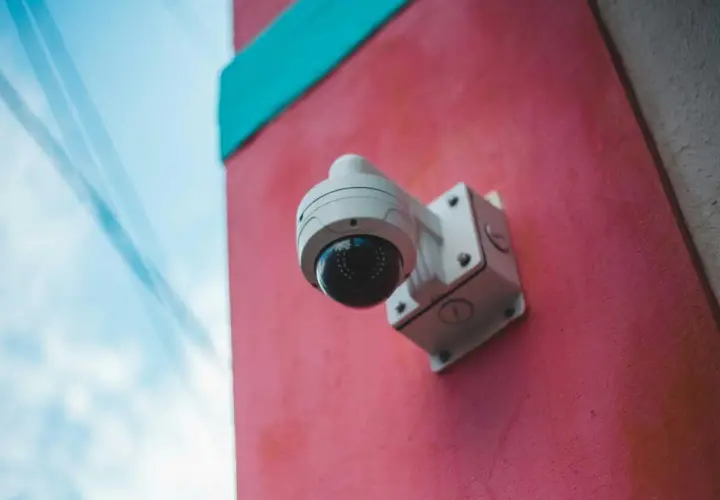 Caméra de surveillance blanche fixée sur un mur rose vif, sous un ciel bleu. Un rappel visuel de la loi sur la caméra de surveillance.